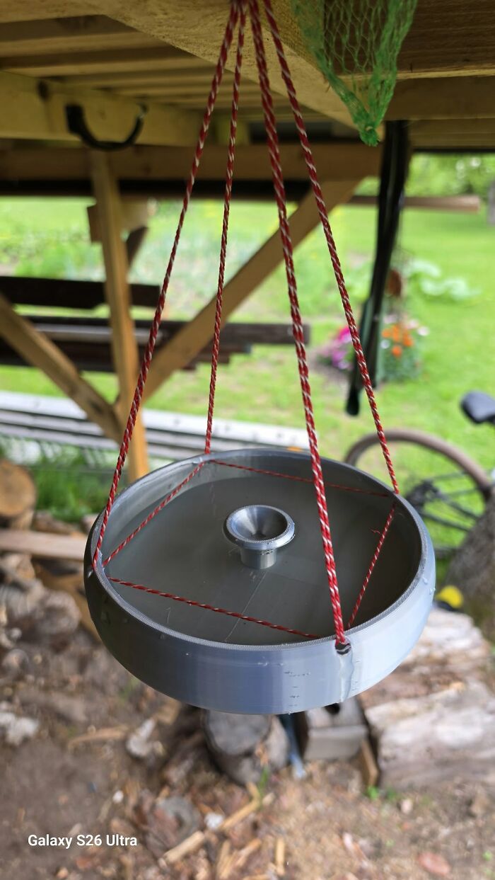 A gray, circular bird feeder with red and white string, hanging outdoors under a wooden structure, a functional thing created using a 3D printer.