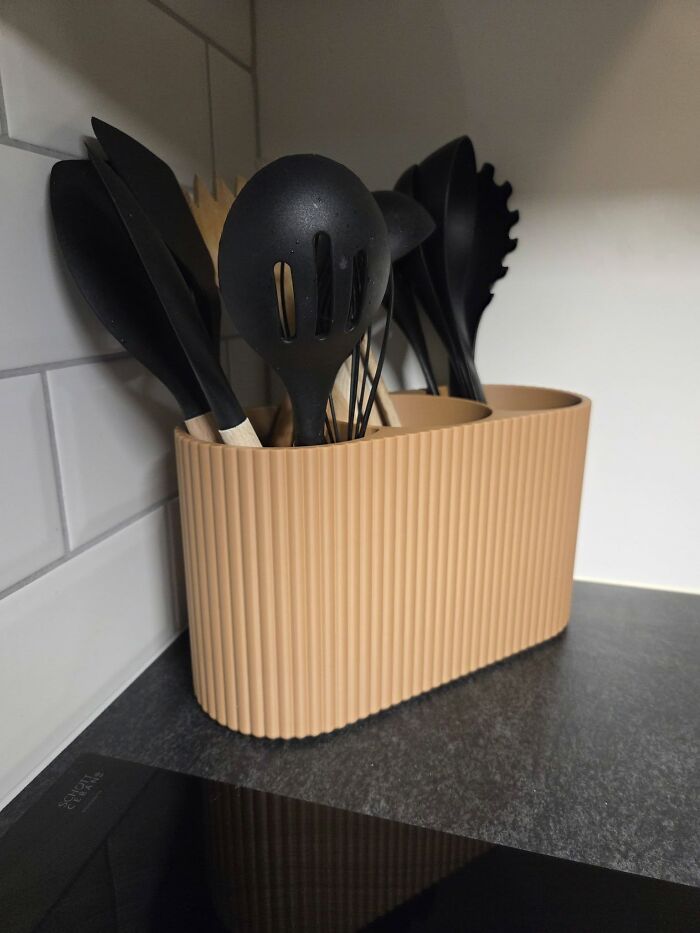 A modern kitchen utensil holder, 3D printed in a ribbed design, stands on a dark countertop.