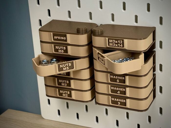 Two stacks of brown, 3D printer created storage drawers, labeled with nuts and washers, hanging on a white pegboard.