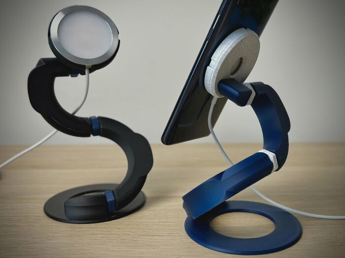 A pair of 3D printer created smartphone stands, one black and one blue, holding phones with chargers.