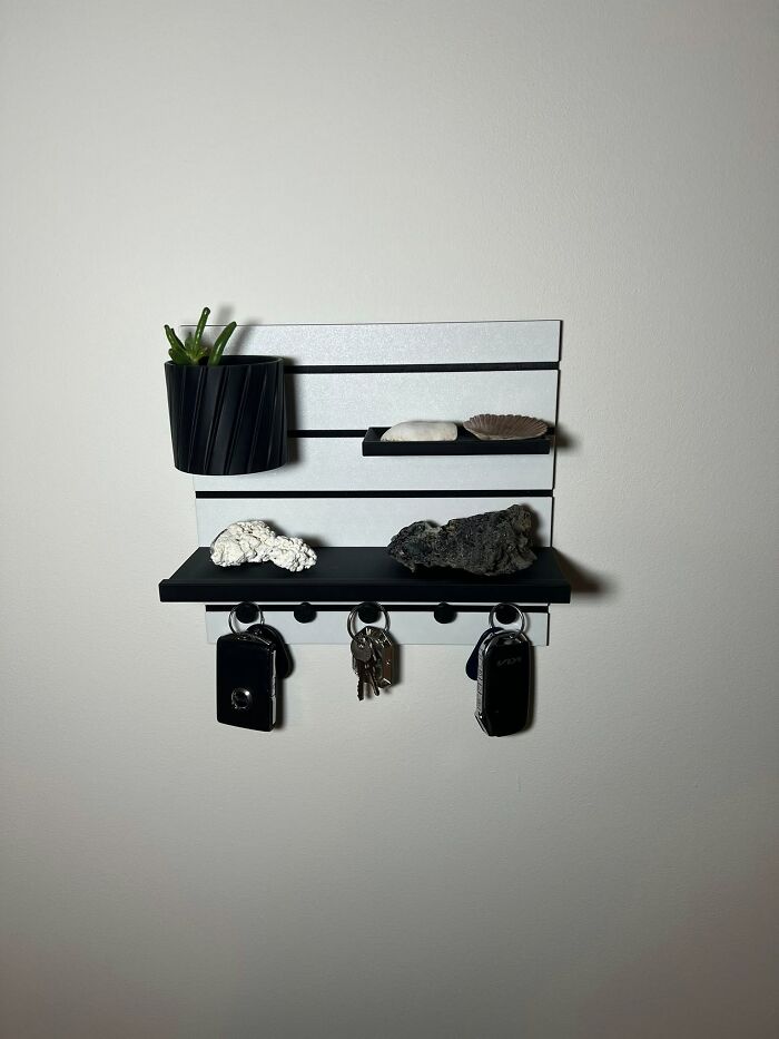 A modern 3D printed wall organizer with shelves and hooks holds keys, a small plant, and decorative rocks.