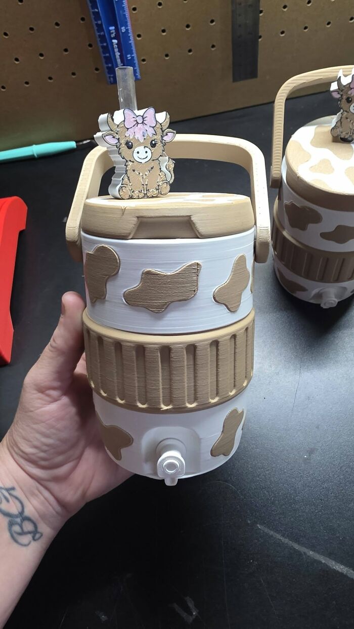 A hand holds a custom 3D printed water cooler with a playful cow pattern and a cute cow figurine on top.