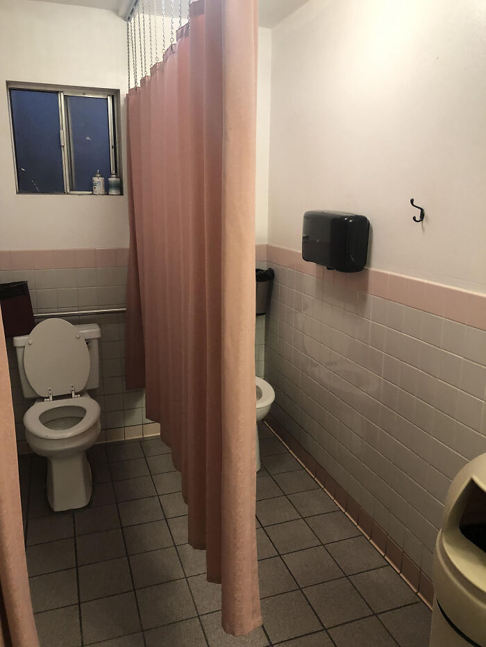 A terribly designed public bathroom with two toilets separated by a hanging pink curtain, and a small window.