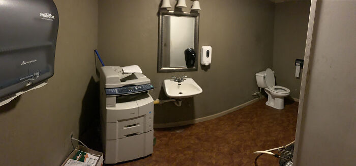 A terribly designed bathroom also serving as an office supply room, featuring a toilet, sink, and a large copier machine.