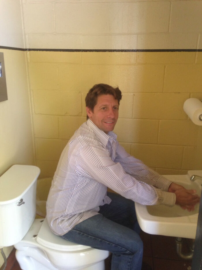 Man sitting on a toilet while washing hands in a sink, highlighting a terribly designed bathroom layout.