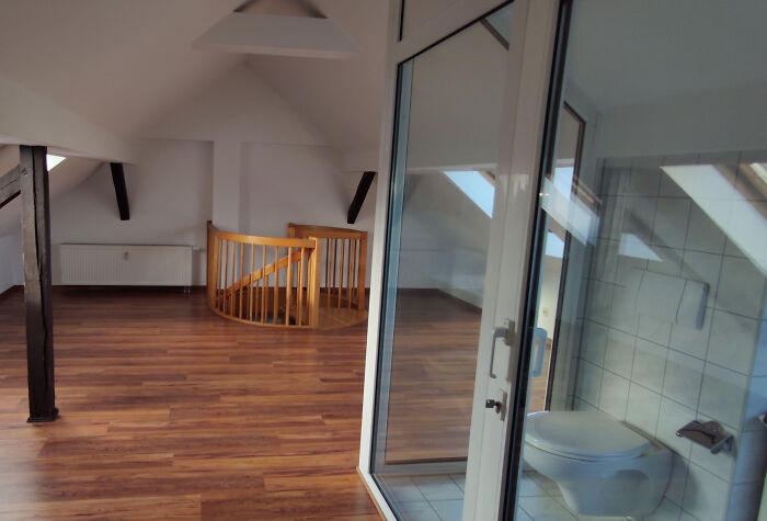 A terribly designed bathroom with a toilet visible through a glass door, situated in a large open room.