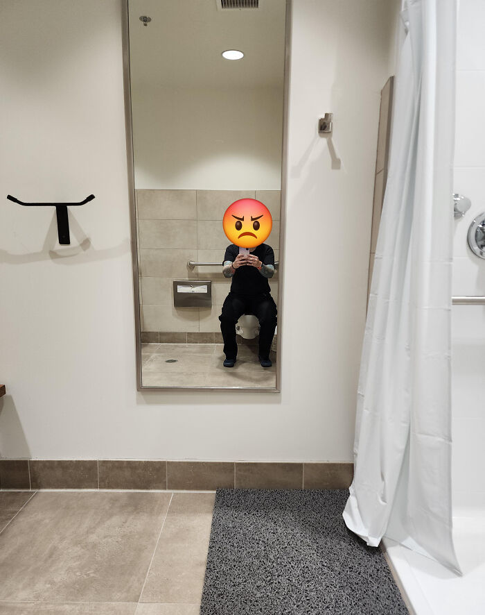 A reflection of a person with an angry emoji face sitting on a toilet in a terribly designed bathroom.