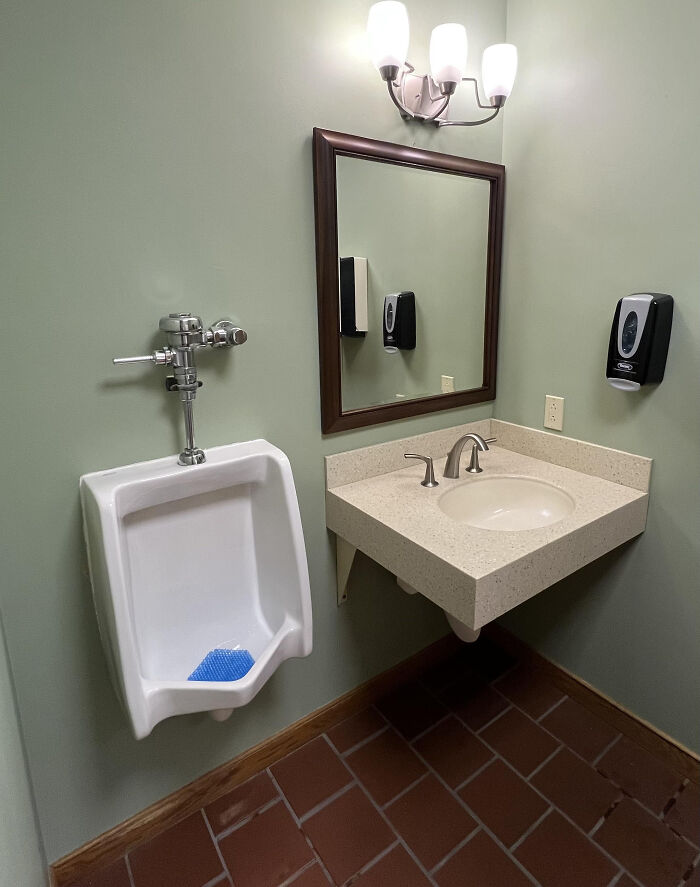 A terribly designed bathroom featuring a urinal next to a sink, with green walls and terracotta floor tiles.