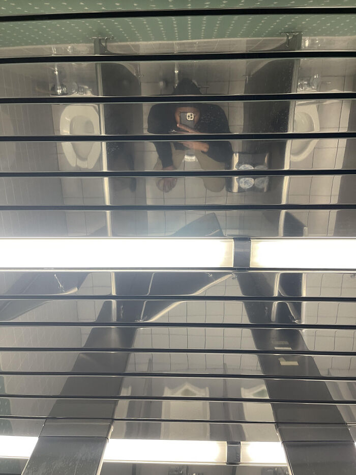 An upward shot reflecting a person holding a phone and two toilets in a highly reflective ceiling, a terribly designed bathroom.