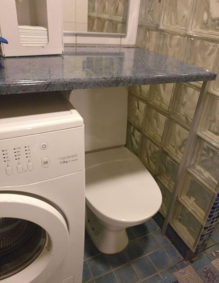 A terribly designed bathroom with a toilet squeezed between a washing machine and a glass block wall.