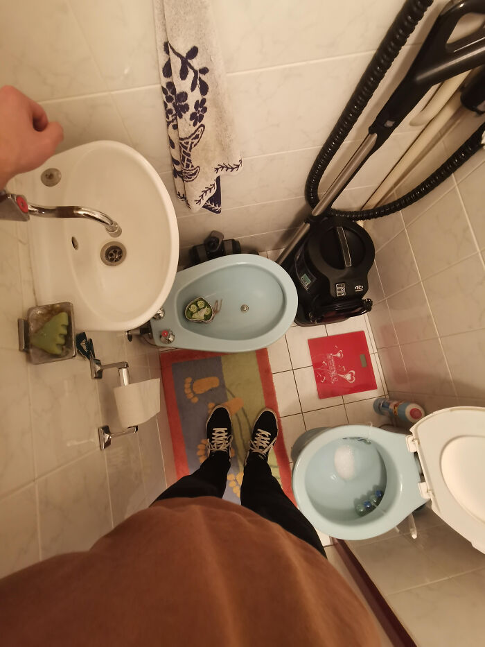A top-down view of a small, terribly designed bathroom with a person's feet, a toilet, bidet, sink, and vacuum.