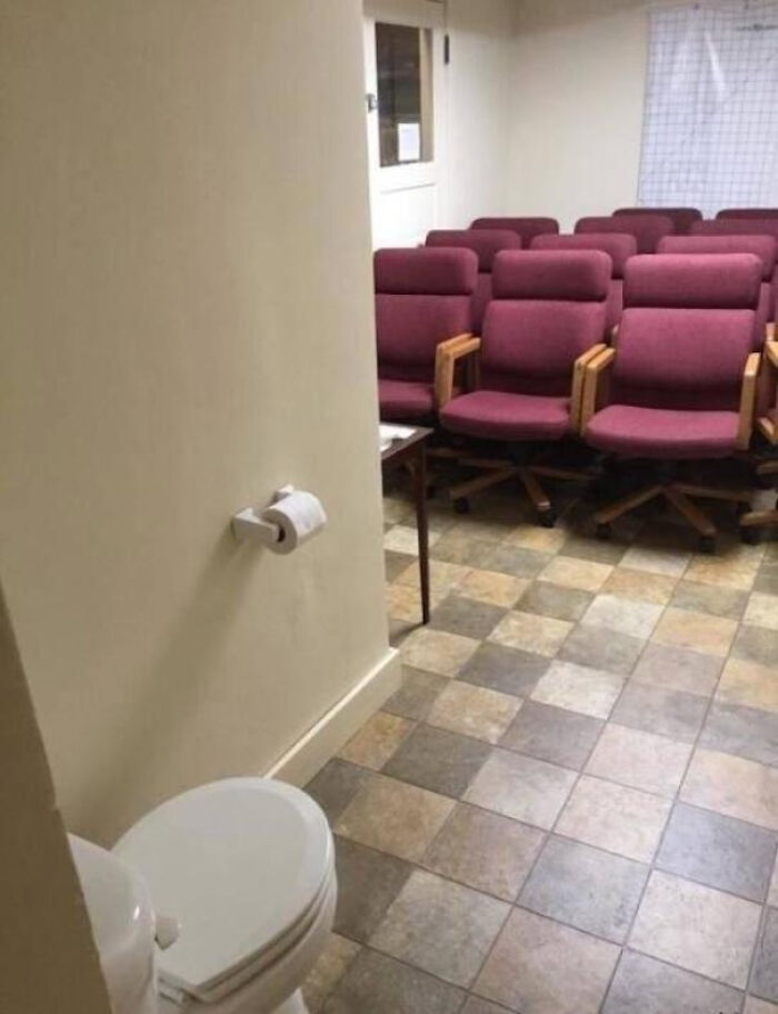 A terribly designed bathroom with a toilet openly placed next to an office meeting room with red chairs.