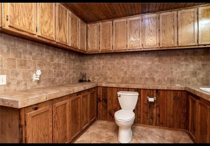 A terribly designed bathroom featuring a toilet in the middle of a kitchen-like area, surrounded by wooden cabinets and counters.