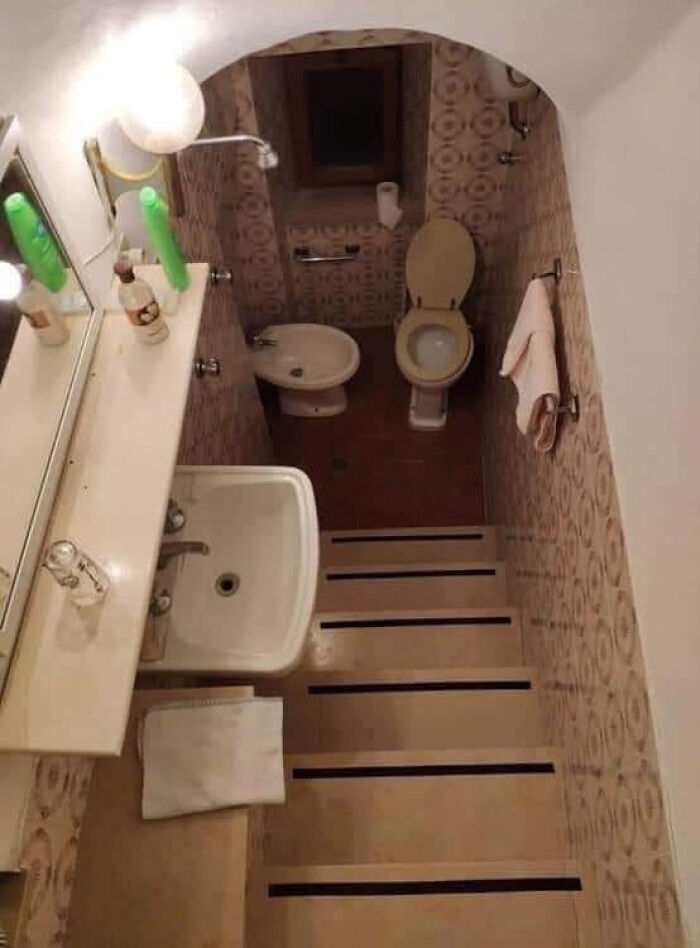 A terribly designed bathroom with a toilet and bidet at the bottom of a steep, narrow set of stairs, next to a sink.