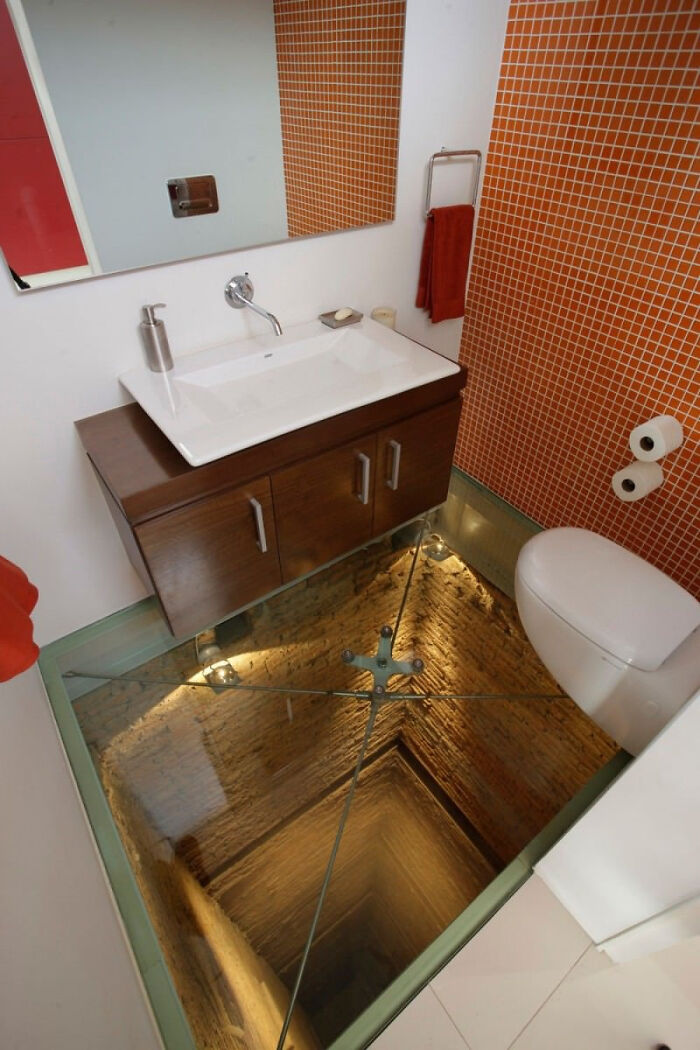 A terribly designed bathroom with a toilet and sink over a glass floor looking down into a deep well.
