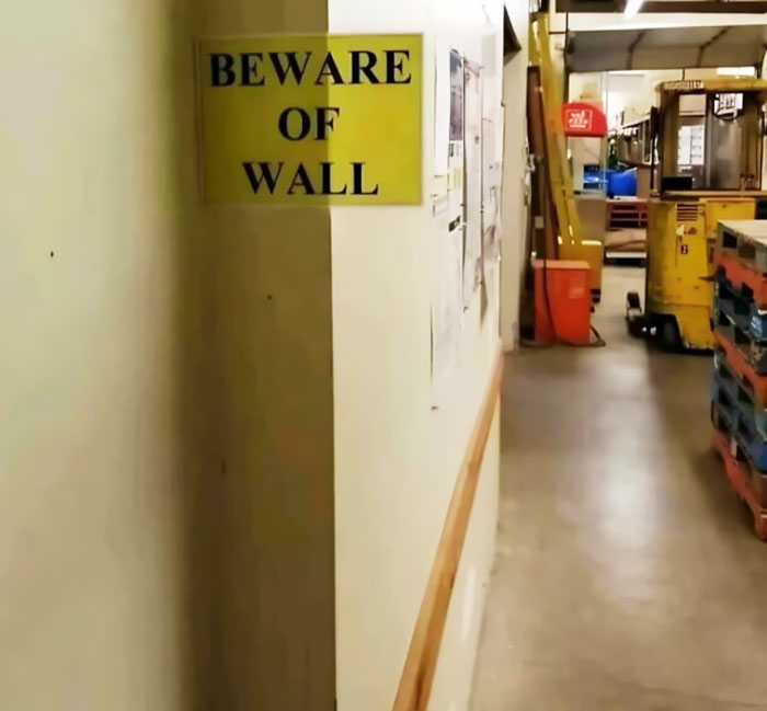 A hilariously failed sign says Beware of Wall on a blank wall in a warehouse.