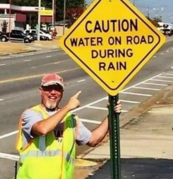 A man in a neon vest hilariously failed holding a sign that reads CAUTION WATER ON ROAD DURING RAIN on a sunny day.