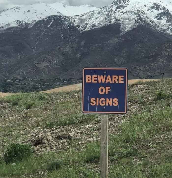 A hilariously failed sign in a grassy field with mountains in the background, reading BEWARE OF SIGNS, failing its purpose.