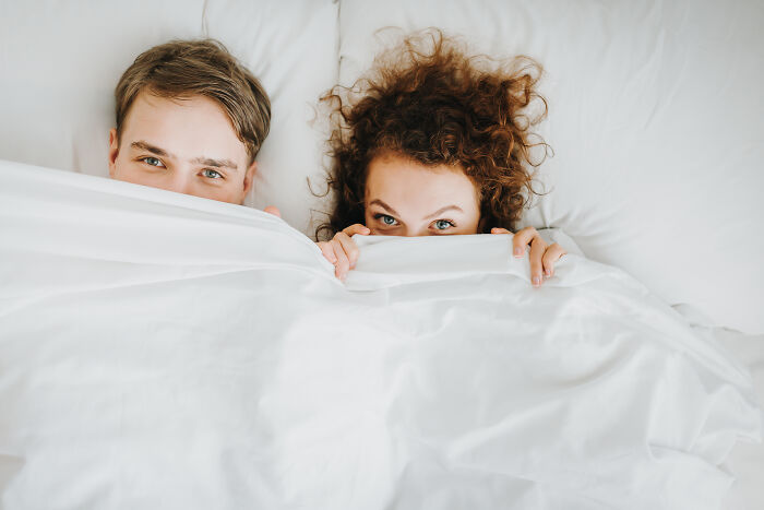 A couple peeking over a white duvet in bed, representing guests who completely ignored basic house etiquette.