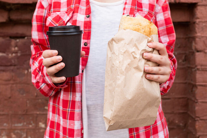 Hands holding a take-out coffee cup and a paper bag with food, showcasing common things we often consume.