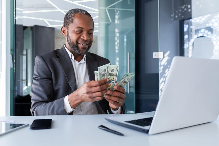 A man happily counting money at a desk with a laptop, symbolizing common things that may turn out to be worse than we realize.