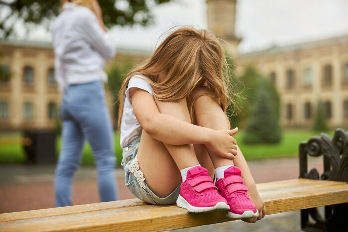 A young girl sitting alone on a bench, looking distressed, conveying a terrifying "we need to leave now" moment.