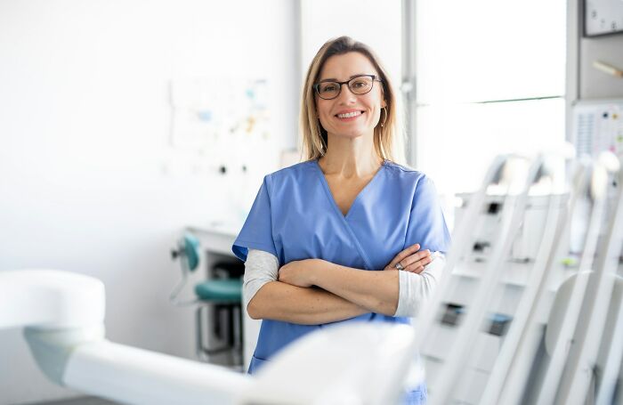 A smiling female doctor in blue scrubs and glasses stands with crossed arms in a brightly lit clinic, showcasing micro feminism clap backs.
