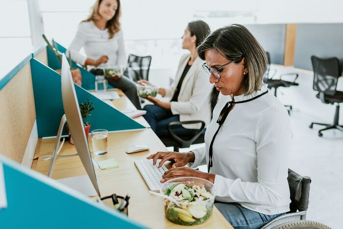 An office worker in a wheelchair types at a computer with a salad nearby, depicting a diverse office story.