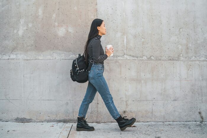A woman in a striped shirt, jeans, and boots walks with a backpack and coffee, embodying micro feminism.