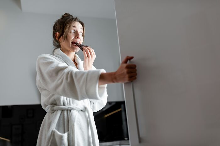 A woman in a white bathrobe secretly eats a chocolate bar, looking surprised. An image related to wedding regrets.
