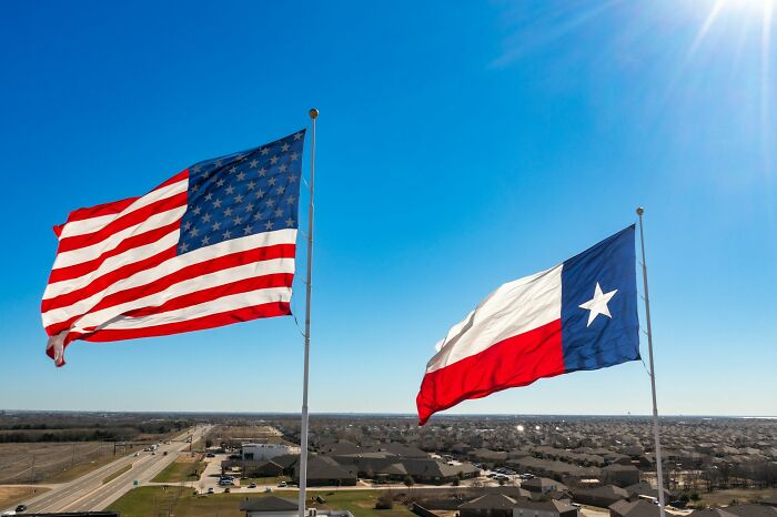 American and Texas flags flying high against a bright blue sky, overlooking a city, reflecting historical myths.