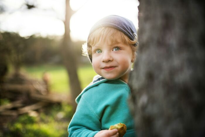 A young child with blue eyes and a cap peeking from behind a tree, relevant to babysitting stories.