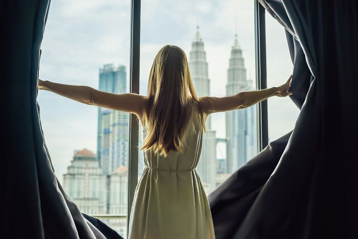 A woman opening curtains to reveal a city skyline with tall buildings. This image reflects unhinged therapist moments.
