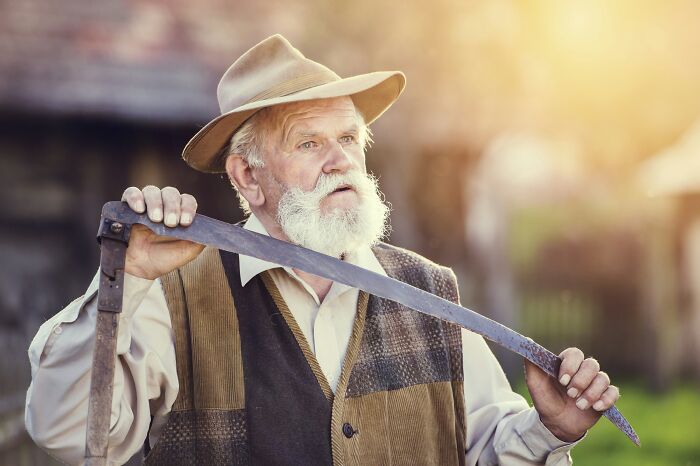 An older man with a white beard and hat holding an old scythe, depicting World War II veterans' extraordinary stories.