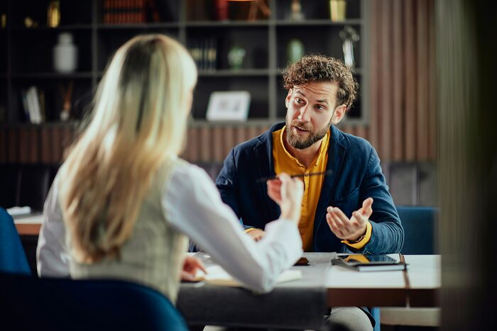 A man with a beard gestures while talking to a woman, depicting a scene with mansplaining examples.