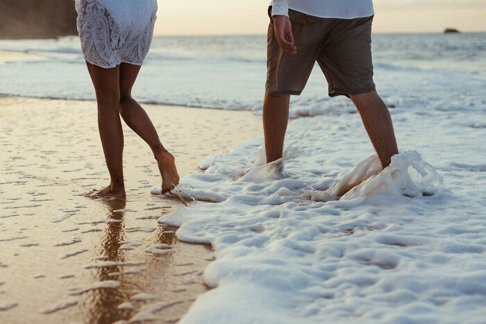 A man and a woman walking barefoot on a beach with ocean waves, hinting at a relationship and being cheated on.