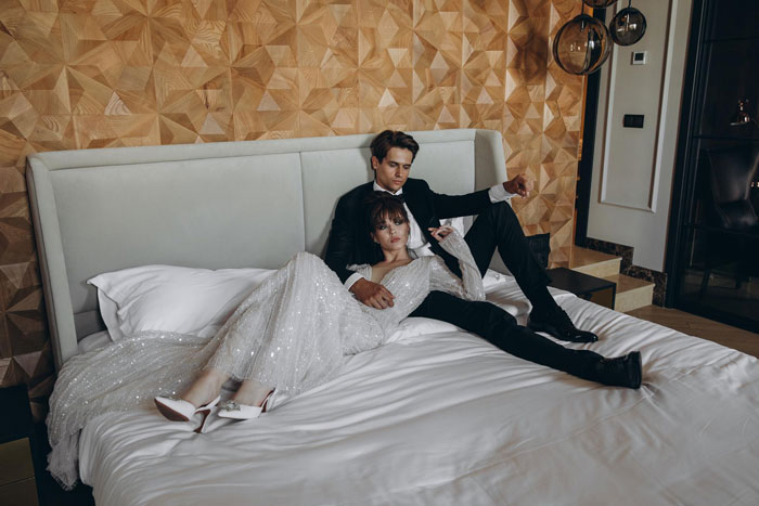 A newlywed couple, the bride in a white dress and groom in a suit, relaxing on their wedding night bed.