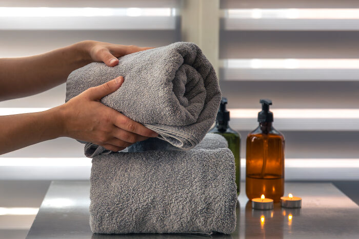 Hands folding gray towels, with candles and soap bottles in the background, contrasting with ignored basic house etiquette.
