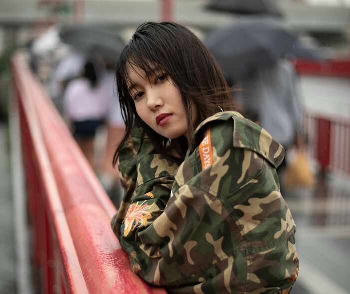 A young woman in a camouflage jacket leans on a red railing, looking at the camera. Products, Trends, and Items.