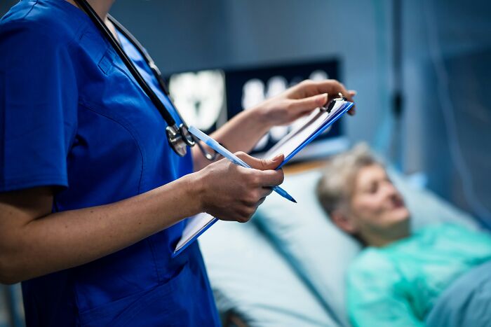 A nurse holding a clipboard stands next to a sleeping patient, hinting at hospital horror stories.