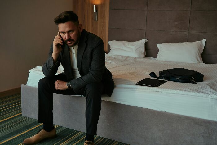 A man sitting on a hotel bed, looking worried while talking on his phone. Uncover hotel industry secrets for your next vacation.