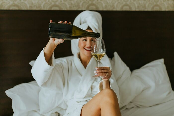 A woman in a bathrobe pours wine in a hotel bed, enjoying vacation comfort and hotel industry secrets.