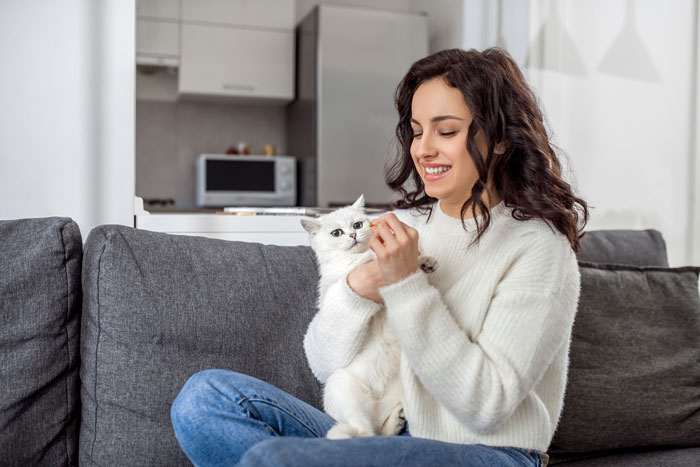 A smiling woman holding a white cat on a couch, after a house sitter caused a crashed car and lost cat incident.