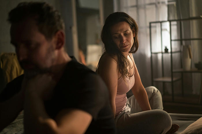 A distressed woman sits on a bed while her husband turns away, deeply affected by old pics with ex and overheard words. A distressed woman sits on a bed while her husband turns away, deeply affected by old pics with ex and overheard words.