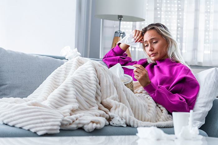 A sick wife, wrapped in a blanket on the couch, looking unwell with tissues around her.