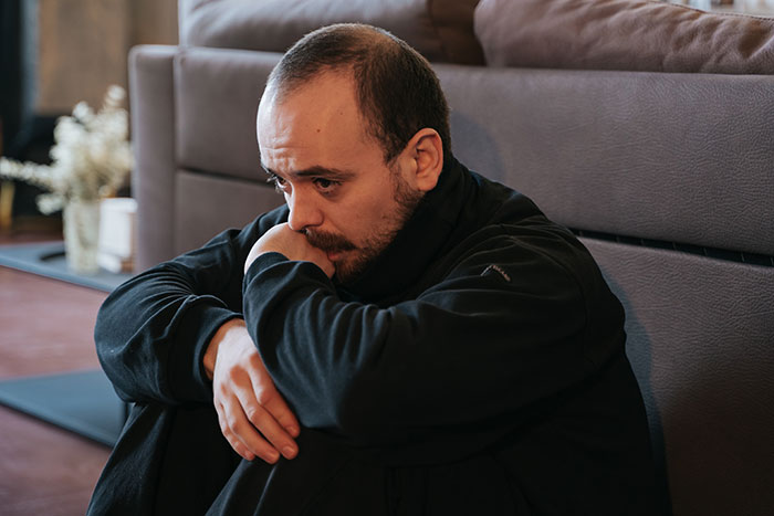 A man sits on the floor, looking pensive and burdened, as he carries the family while his sick wife sleeps. He wonders if he can keep going.