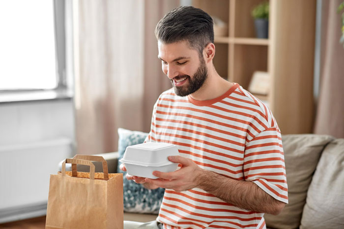 A happy guy unboxing takeout food. A man is smiling, holding a take-out box from a paper bag. A happy guy unboxing takeout food. A man is smiling, holding a take-out box from a paper bag.