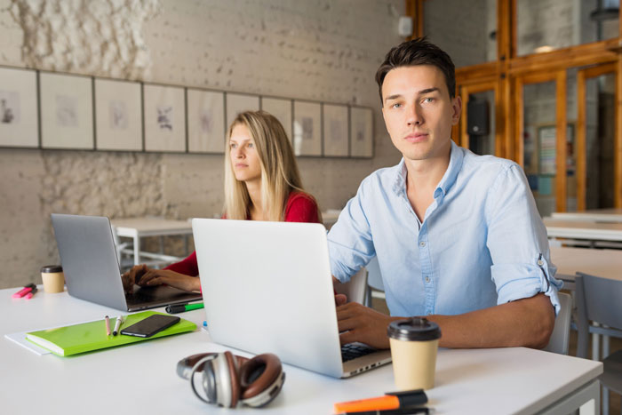 A man, possibly the cheating husband, looks at the camera while a woman, possibly the coworker, works on her laptop in the background. A man, possibly the cheating husband, looks at the camera while a woman, possibly the coworker, works on her laptop in the background.