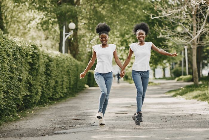Two identical twin sisters joyfully skipping and holding hands in a park, showcasing the chaos and fun of being a twin.