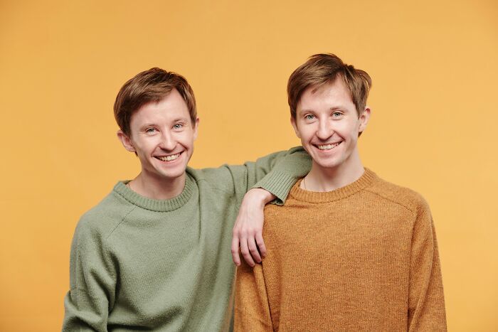 Two smiling identical twin brothers wearing sweaters, one with his arm around the other, against a yellow background, highlighting the chaos of being a twin.